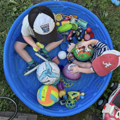 To børn leger med bolde og legetøj i et blåt plastikbassin udenfor.