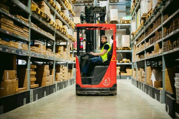 En mand i gul refleksvest sidder i en rød truck på et lager omgivet af høje reoler med kasser