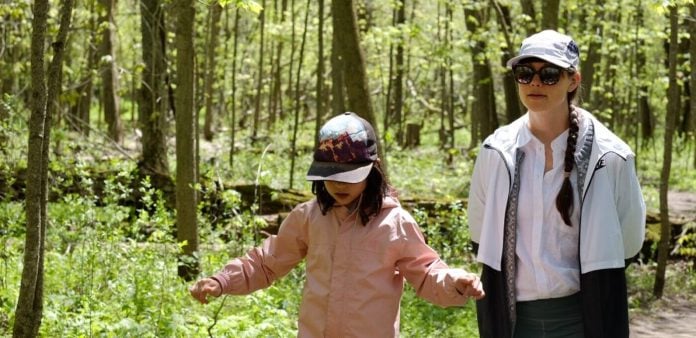 En voksen med mørke solbriller går tud i en skov med en pige med stor kasket til at skærme for lyset
