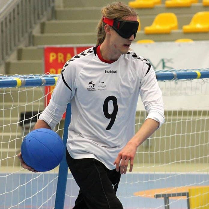 Gustav Koch Rasmussen i en goalball-kamp, hvor hans øjne er blændede, han har en hvid bluse på og en blå bold i hånden